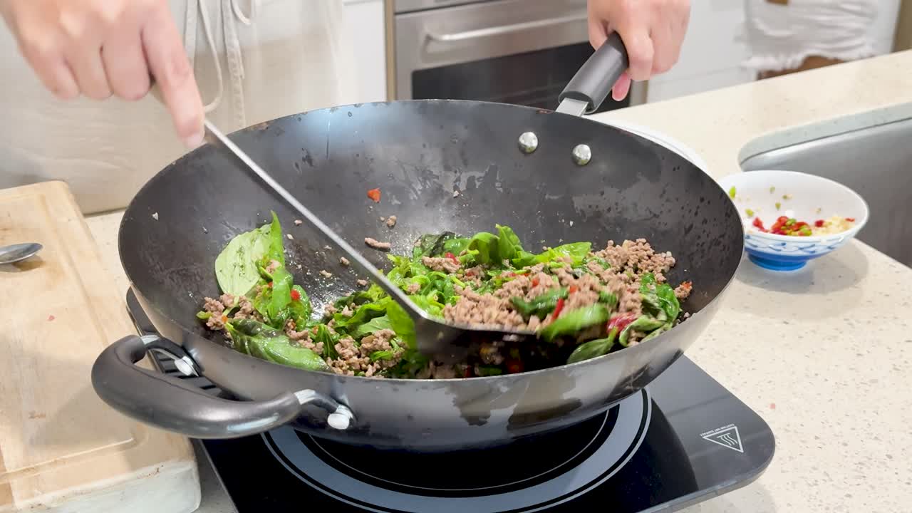 Cook prepares Thai-style stir fry with pork, basil, and vegetables in a hot wok on a modern kitchen countertop with bright, natural lighting