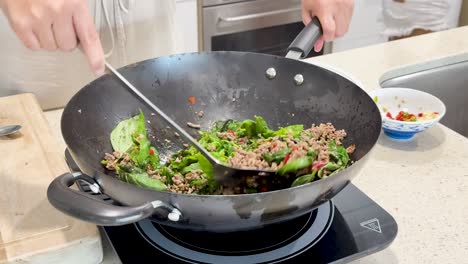 cook prepares thai-style stir fry with pork, basil, and vegetables in a hot wok on a modern kitchen countertop with bright, natural lighting