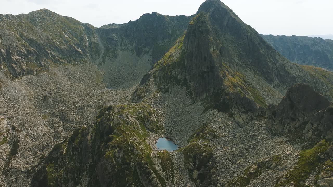 Small glacial blue lake at the bottom of a tall mountain peak, surrounded by mountain ranges and rocks, tracking drone shot