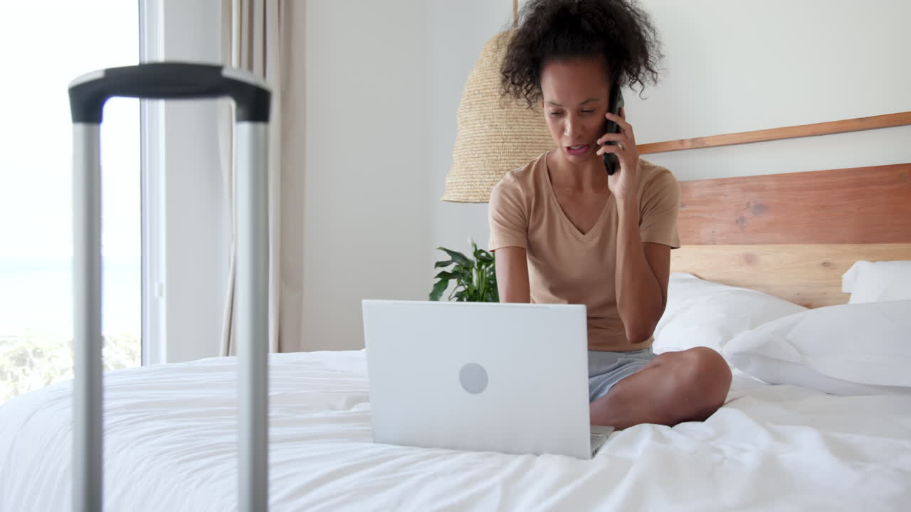 woman sitting on bed using laptop and talking on phone, at home