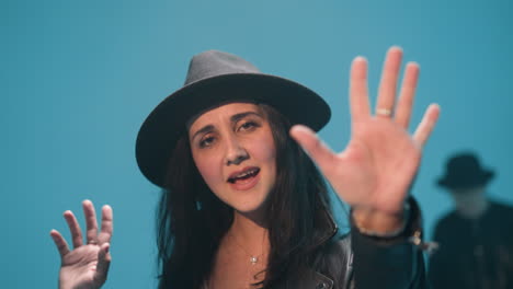 close-up of a lady wearing a black hat and black leather jacket, singing with a serious expression, hands in motion as the camera zooms out. the background is blue