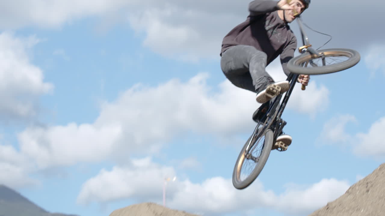 Extreme Sport Mountain Biking. An athletic man on a bike dirt jumping in slow motion.  Blue sky, scattered clouds, dirt jumps and mountains in the background.