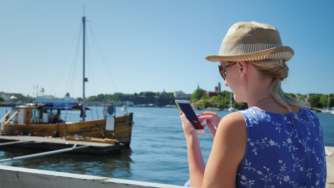 A Resting Woman With A Smartphone Sits On The Embankment Against The Backdrop Of The Sea And Moored 