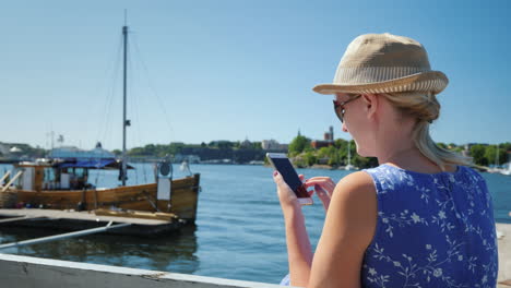 a resting woman with a smartphone sits on the embankment against the backdrop of the sea and moored