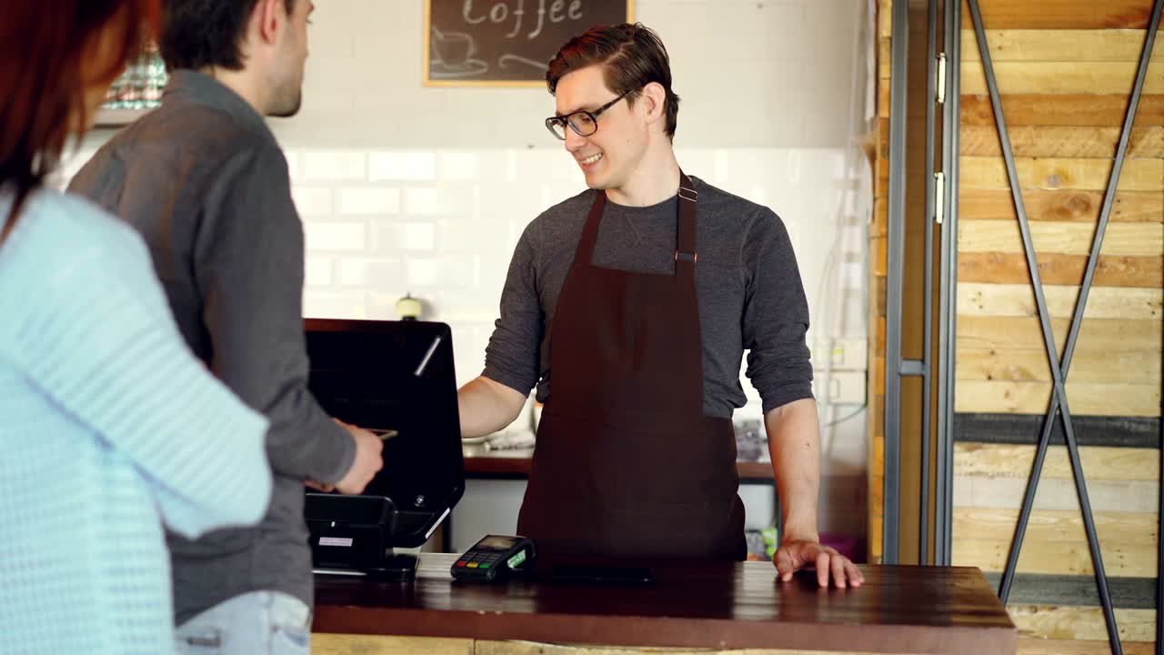 Cheerful friendly cashier is taking orders from customers standing in line, accepting contactless payment with smart phone and selling takeaway coffee.