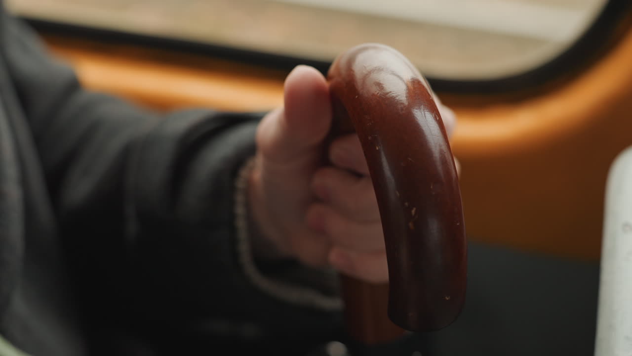 Primo piano di una mano che afferra la maniglia di legno all'interno di un autobus, il creatore che regola la presa mentre il polsino del cappotto scuro sfiora il finestrino, texture lucida e umida sulla maniglia, dita che si incurvano in un gesto di pendolarismo costante, umore riflessivo.