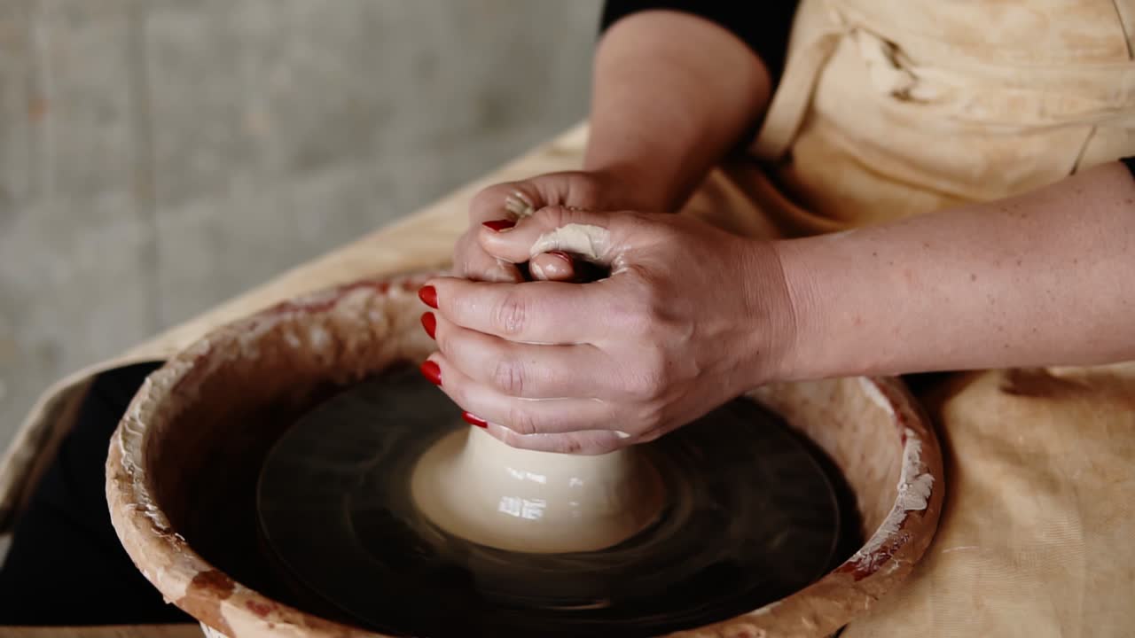 Close-up of potter's hands with red manicure working with wet clay on a pottery wheel making a clay product in a workshop. Unrecognizable female person