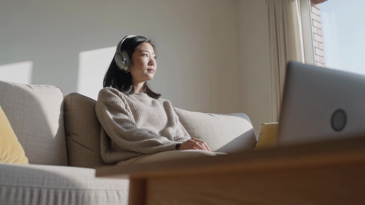 穿着米色毛衣的女子戴着耳机坐在沙发上，头随着阳光斑驳处转动，凝视窗外。