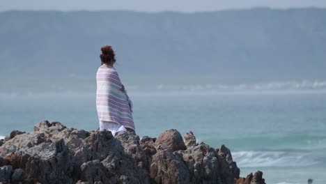 Woman-standing-on-the-shore-looking-over-the-ocean-with-a-towel-around-her