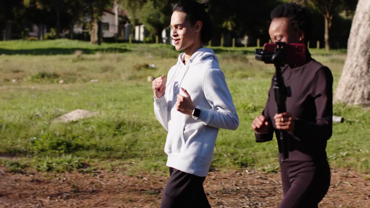 Amici di diverse etnie che fanno jogging motivati dalla luce del sole mattutina, filmando con uno stabilizzatore per telefono lungo un sentiero pavimentato.