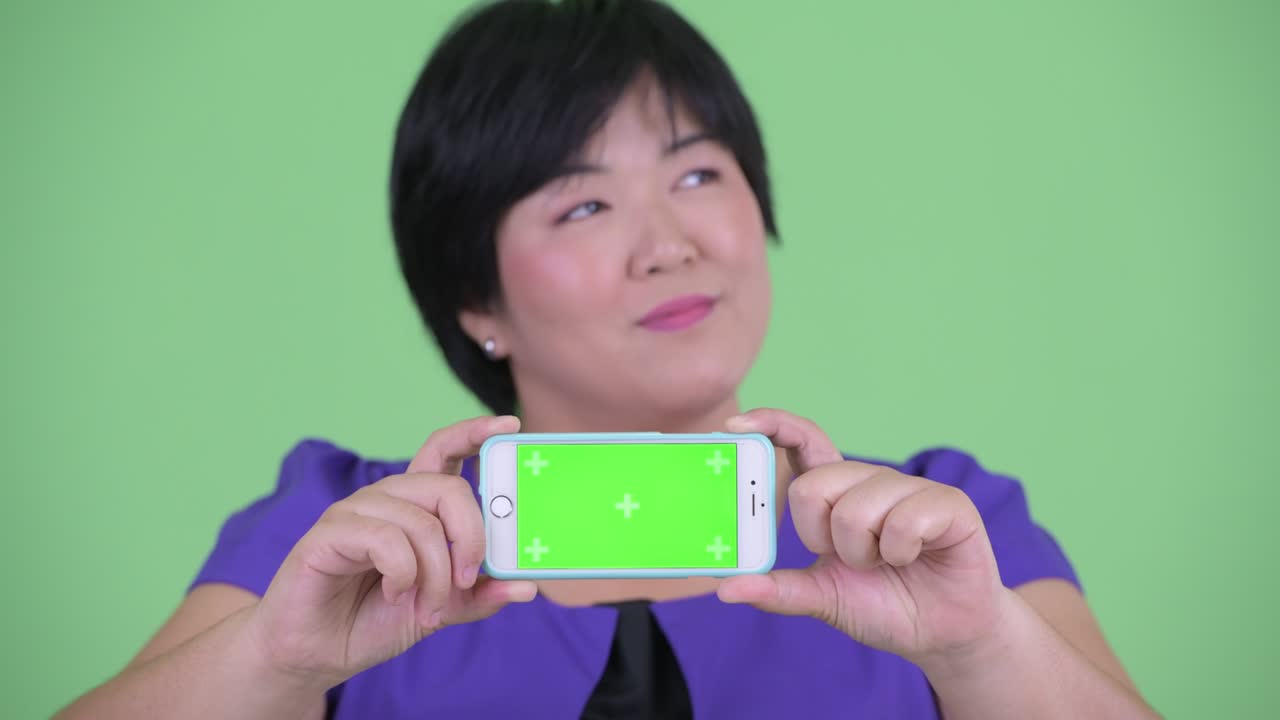 초록색 배경에 휴대 전화에서 크로마 키를 보여주는 짧은 헤어 스타일의 과체중 아시아 여성의 초상화