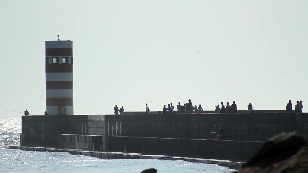 Silhouette of a lighthouse getting hit by big waves while tourists are walking along.