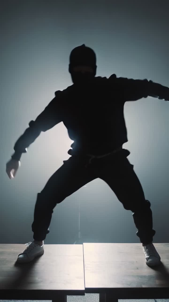 Video verticale: Uomo che balla con felpa con cappuccio, appoggiato al bordo del palco, saltellando e creando un'ombra retroilluminata.