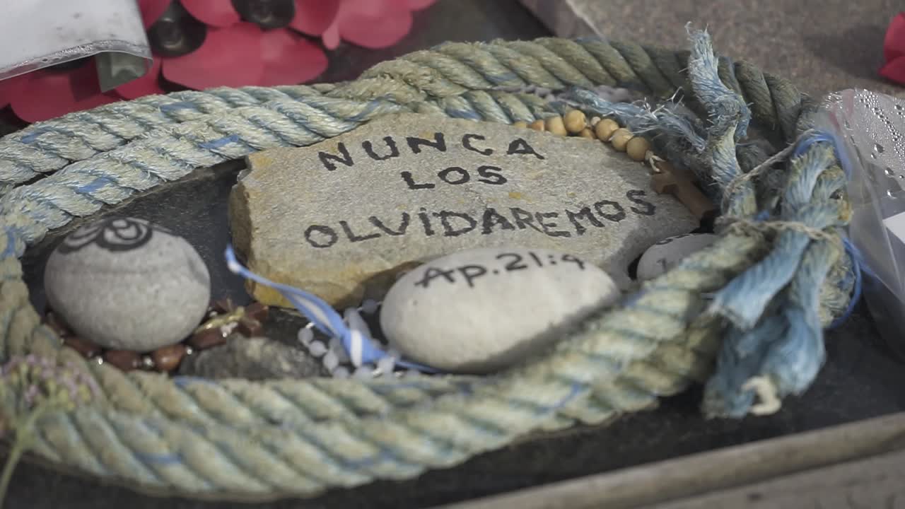 Close-up details of a memorial with the spanish inscription nunca los olvidaremos (we will never forget) alongside rope, stones, and red petals