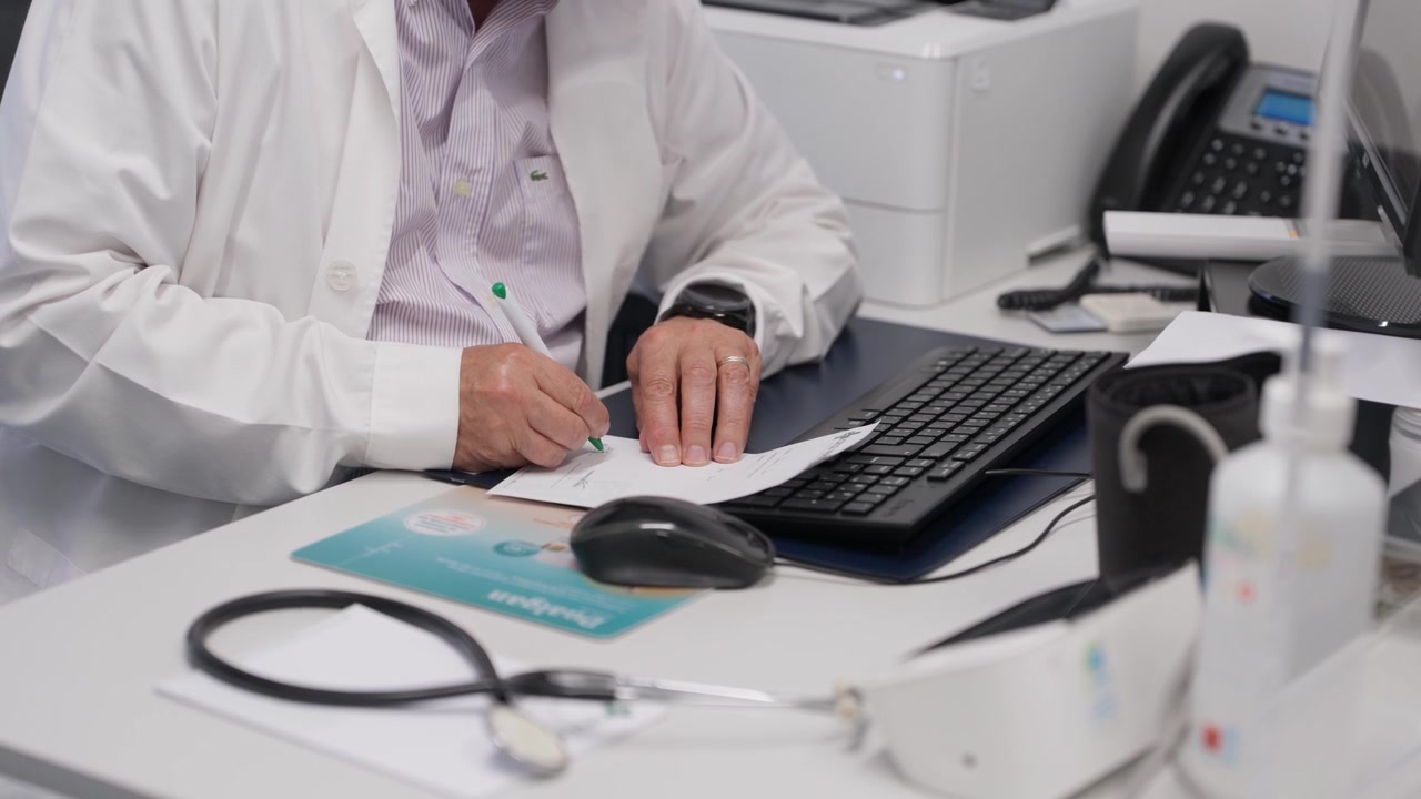 Médico escrevendo em papel em uma mesa com teclado, estetoscópio e mouse de computador.