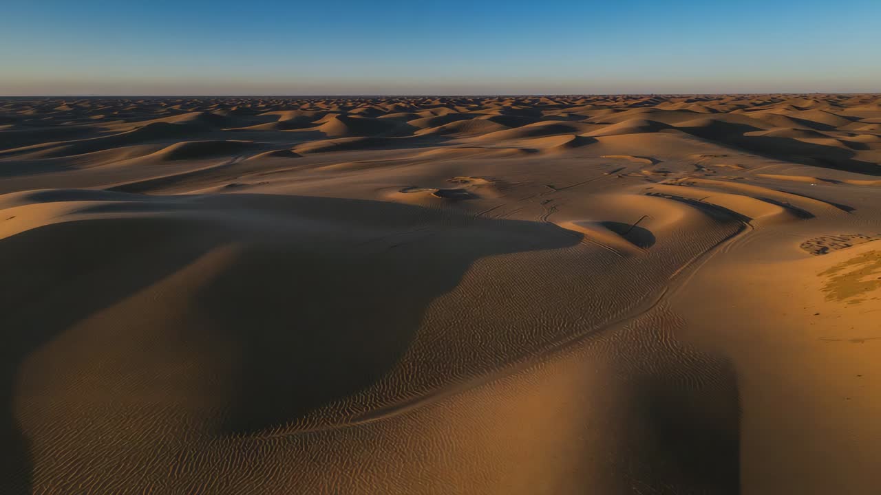 Avvio della camera con ripresa panoramica di un ampio campo di dune per evidenziarne i contorni, rivelando increspature e tracce di pneumatici