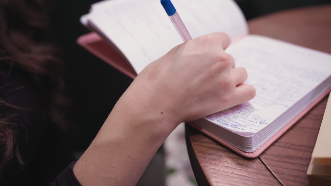 Close up of light skin hand holding blue pen writing on open book placed on wooden table, detailed handwriting fills lined pages, warm indoor setting enhances focus and calm atmosphere