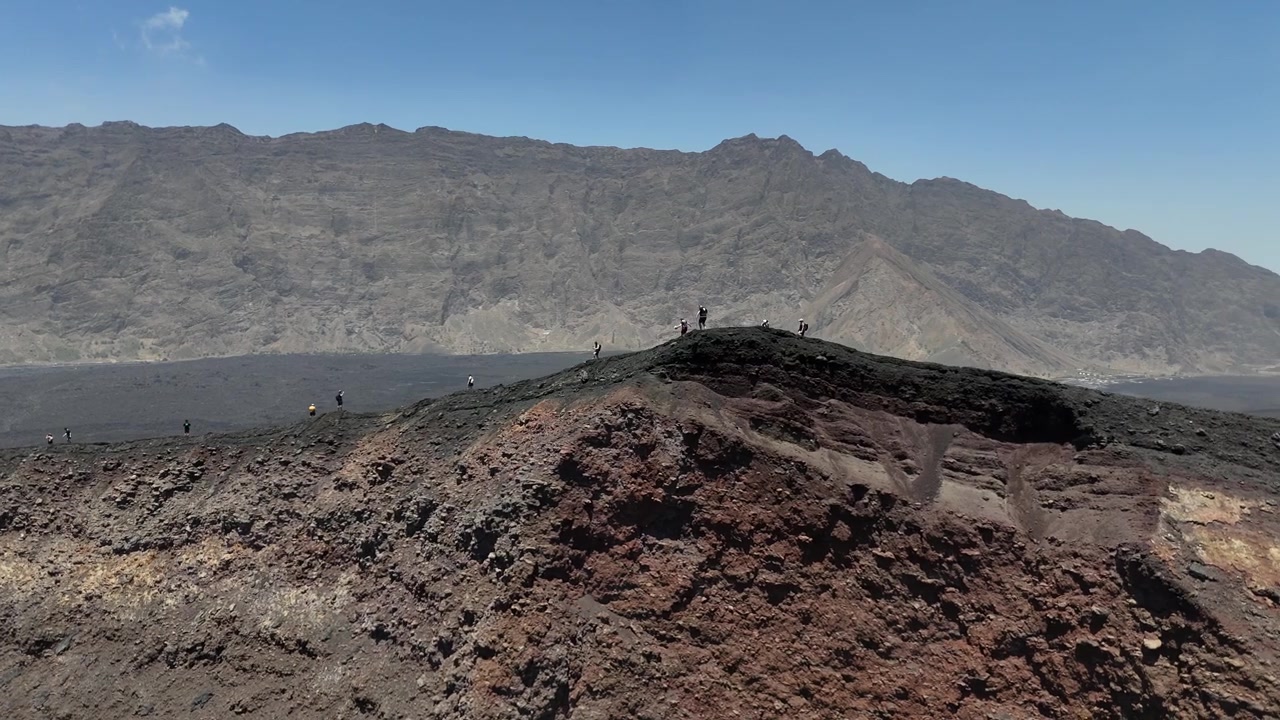 Ripresa con drone di escursionisti che fanno trekking sul bordo frastagliato del cratere del Pico do Fogo, Capo Verde, con la spettacolare parete della Bordeira e le pianure vulcaniche di Chã das Caldeiras sullo sfondo.