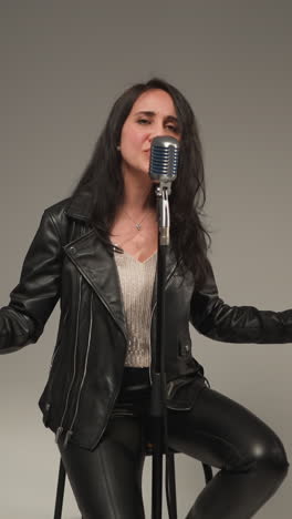vocalista con una chaqueta de cuero negra sonriendo y cantando con manos expresivas, ojos abiertos, sentado en un taburete alto, guitarrista tocando una guitarra roja, tecladista tocando un sampler rojo sobre un fondo gris