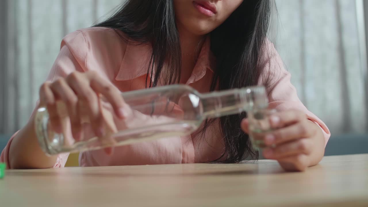 술에 취한 우울한 아시아 여성이 술을 마시기 전에 글라스에 보드카를 부어 집에서 울고 있습니다.