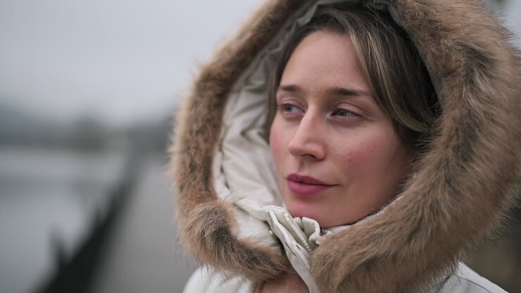 Una donna in piedi vicino all'acqua, che accoglie l'atmosfera fredda con un dolce sorriso. Il suo caldo cappotto con cappuccio di pelliccia la tiene al caldo mentre ammira il paesaggio circostante.