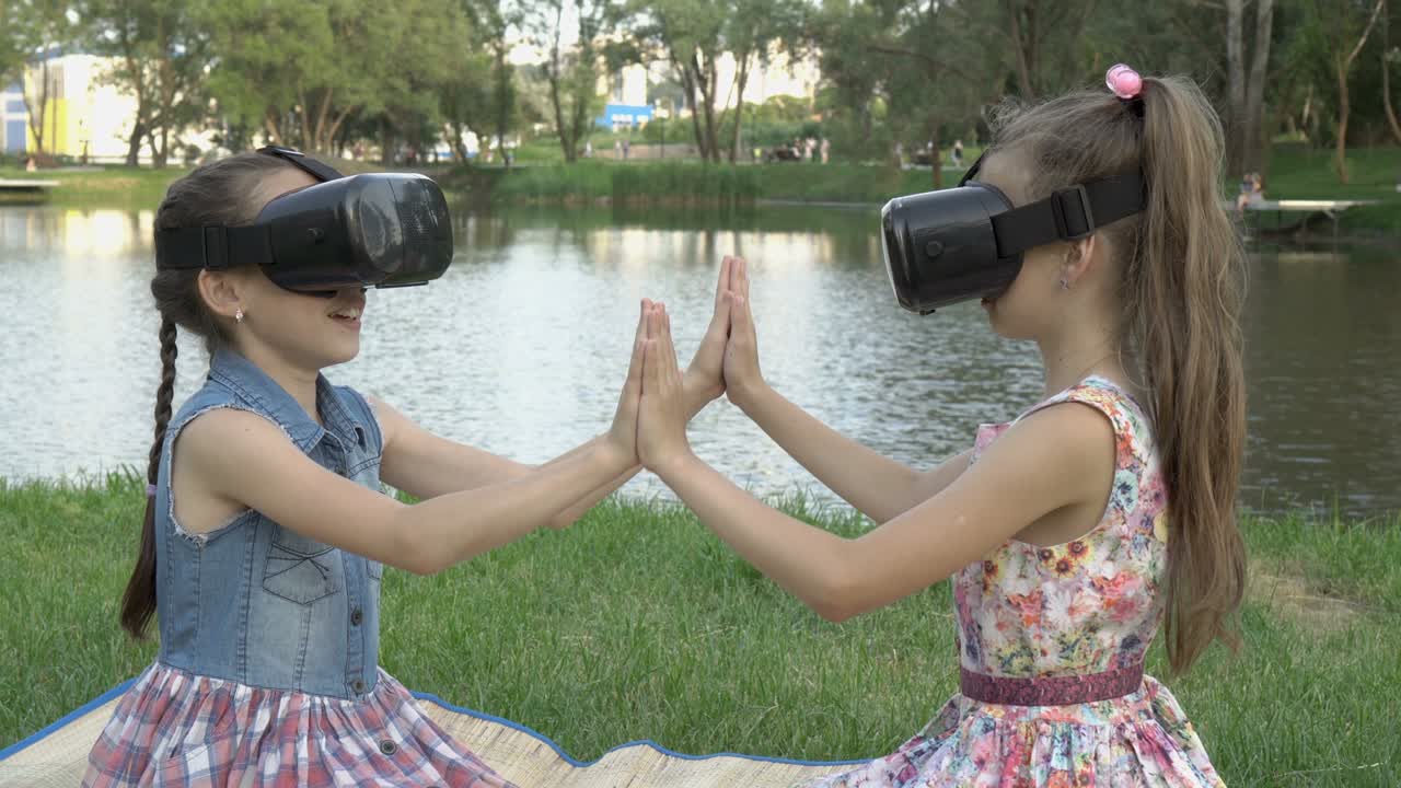 두 어린 소녀는 가상 현실 안경으로 놀고, 여름 저녁에 물을 배경으로 공원에서 앉아 서로 손을 잡고 미소 짓고 있습니다.