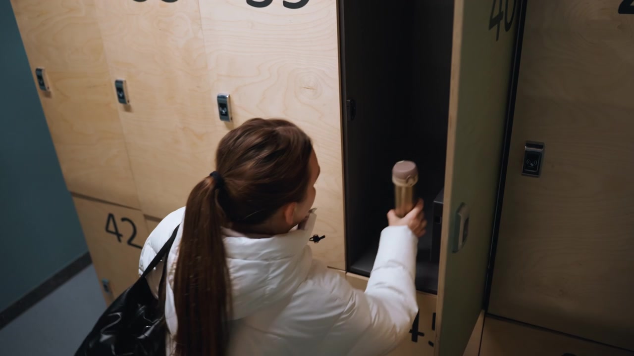 Woman at locker retrieving bottle, insulated thermos pulled from numbered wooden compartment, quick focused motion, keycard access and lock check, energetic preparation for training or work shift