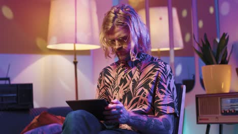 young stylish caucasian man with long hair in headphones sitting on a chair and using tablet in a retro party at home