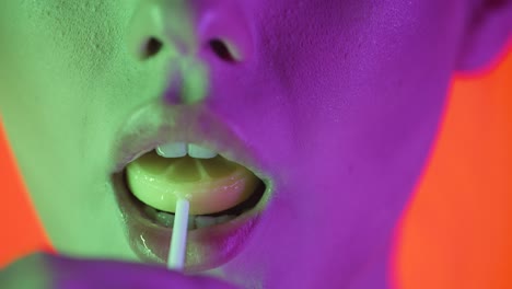 close up shot of young sexy woman lips while she licks a sweet lollipop with pleasure with her tongue against orange background with green purple contrast on face in slow motion