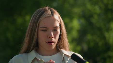 lady with blonde hair stands in outdoor setting, holding green air pump handle with look of surprise, her face shows a curious expression as she examines the handle closely, surrounded by trees