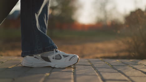 a close-up shot of a person wearing jeans and white shoes, walking slowly on a paved path in a park. the warm, soft light and blurred background