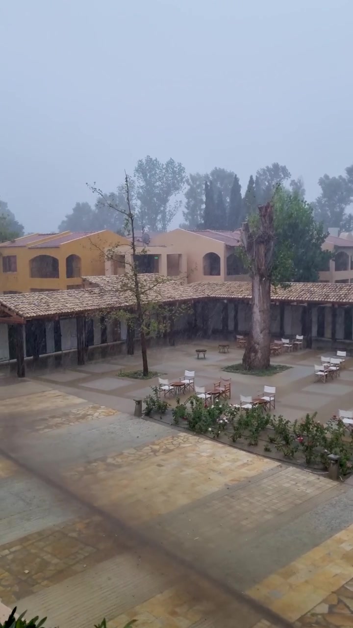 Forte temporale estivo con acquazzone su un cortile di un tradizionale hotel resort mediterraneo a Corfù, Grecia. Elementi meteorologici di una giornata piovosa e tavoli e sedie vuoti di un ristorante/caffè all'aperto.