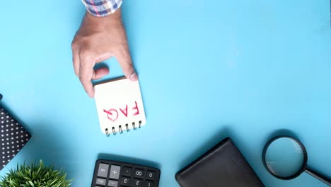 hand holding a notepad with faq written on it, along with other office supplies on a blue surface.