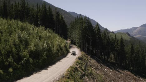 aerial dolly out of a jeep driving on a hillside road surrounded by dense green pine woods, mountains in background, british columbia, canada