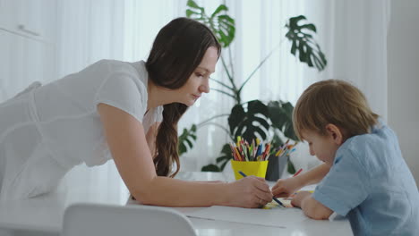 mamá ayuda a su hijo a realizar tareas preescolares para dibujar un dibujo a lápiz sentado en la mesa en la casa