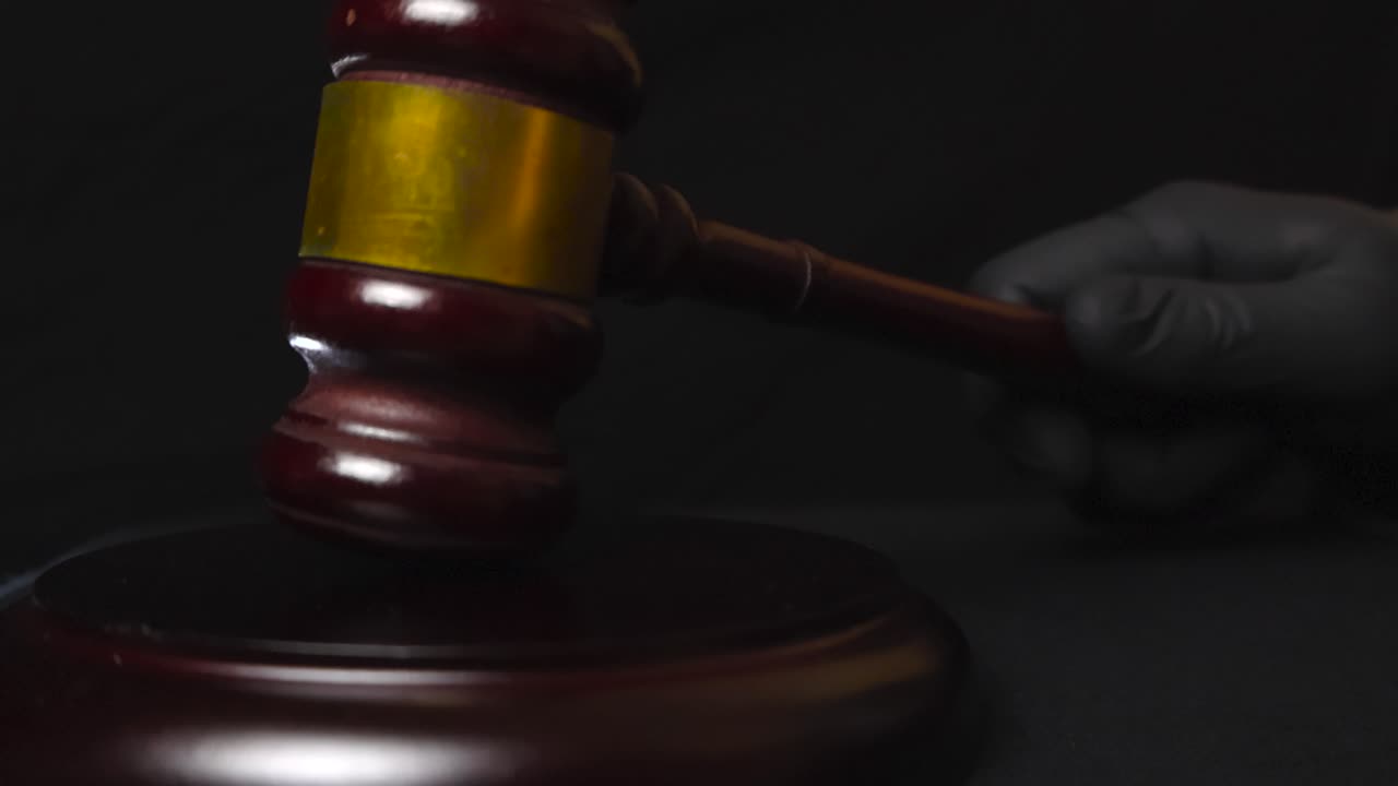 Close up video of a law and justice brown colored wooden court hammer picked up by a gloved black hand and slammed and hit on the table in slow motion twice in front of a black background