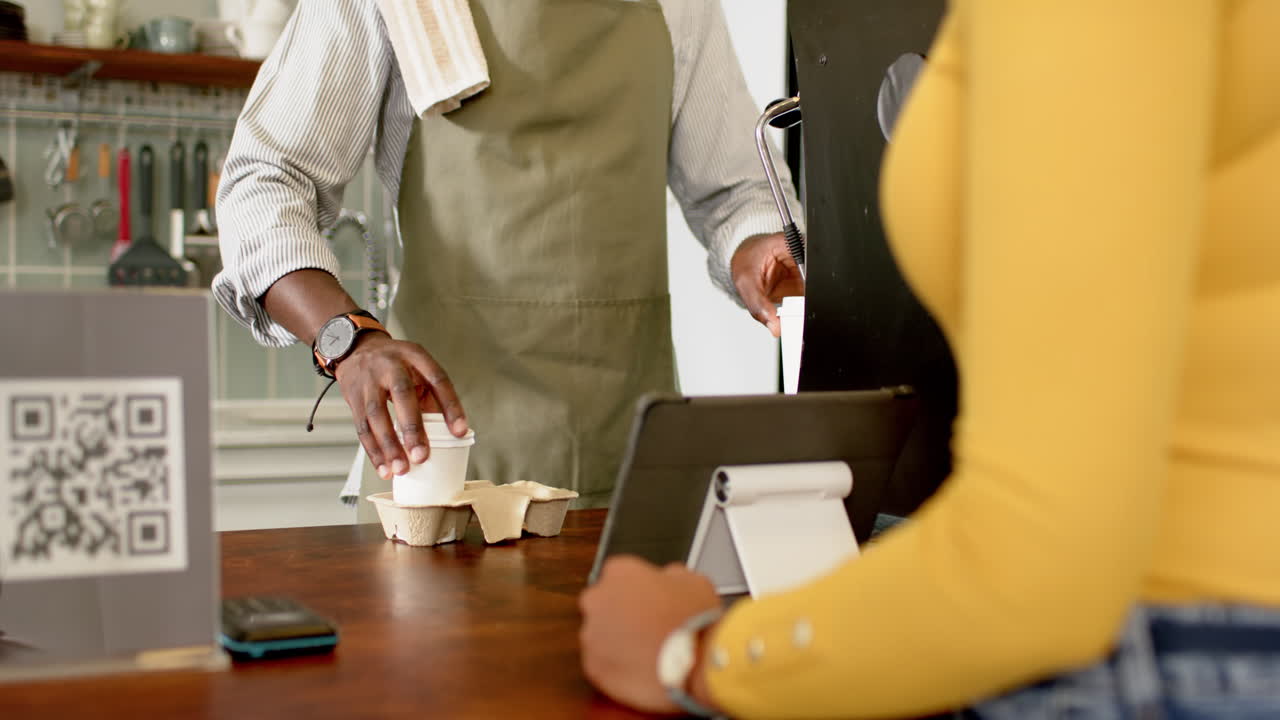 A young African American man serves coffee, standing across a biracial woman with a tablet