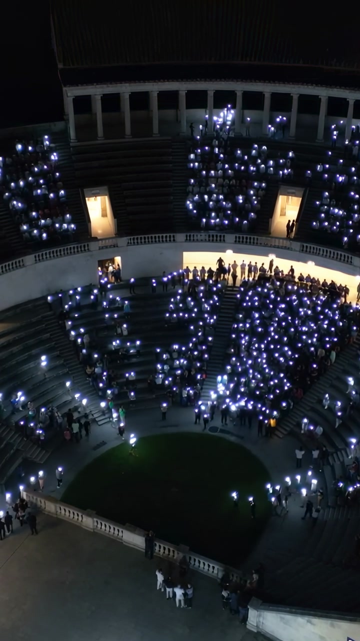 Vertical video: Raising mobile lights, seated crowd shifting in night arena with lit doorways