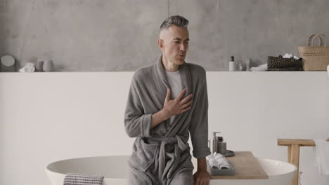 suffering man in pajamas and robe having a chest pain and touching his heart area in the bathroom at home