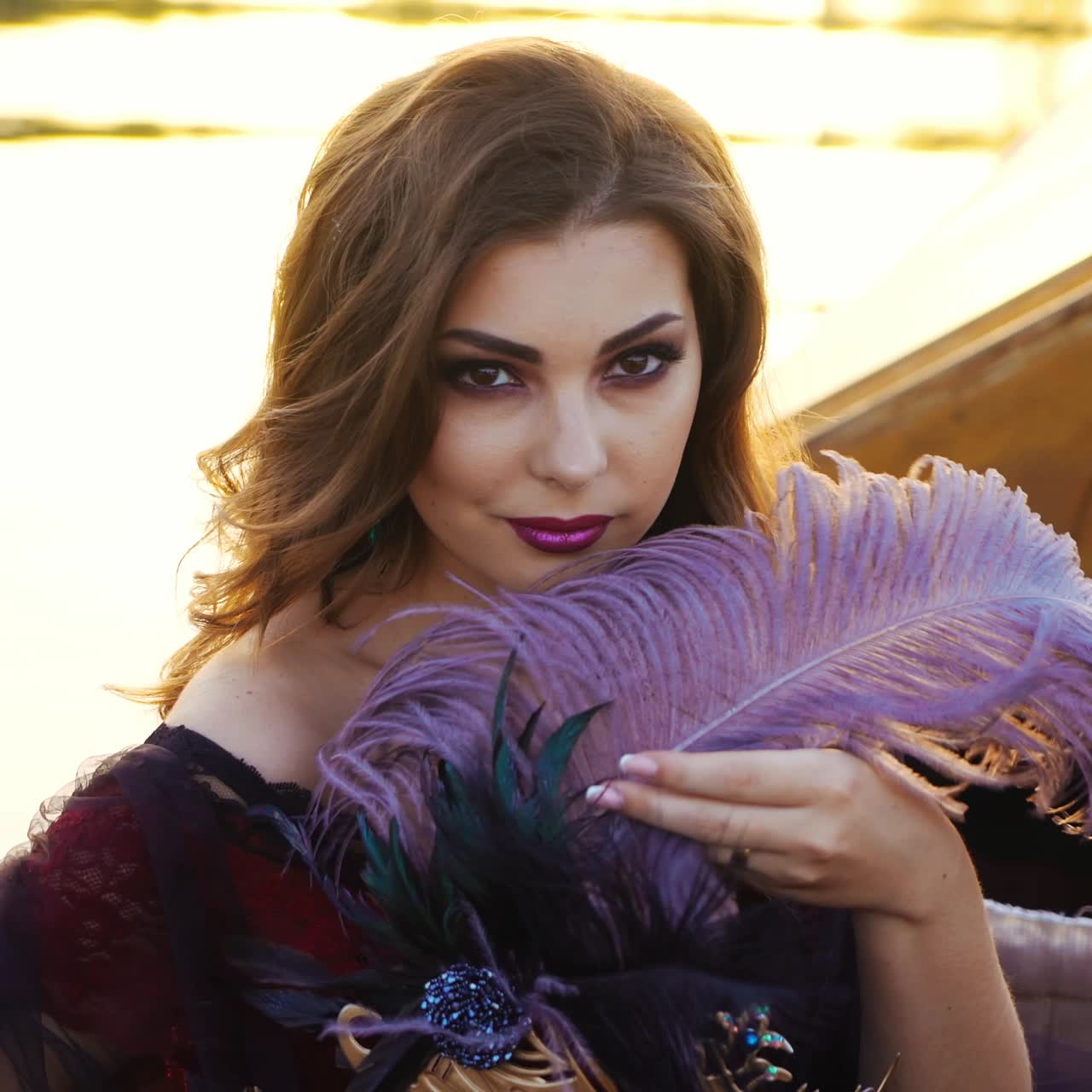 A beautiful woman is sitting in the gondola and touchhing a purple feather with a carnival mask on a blurred background of sunset