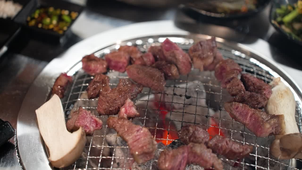 Close-up shot of tongs flipping multiple small, sizzling pieces of Hanu Korean beef and king oyster mushroom on a wire rack over hot glowing charcoal