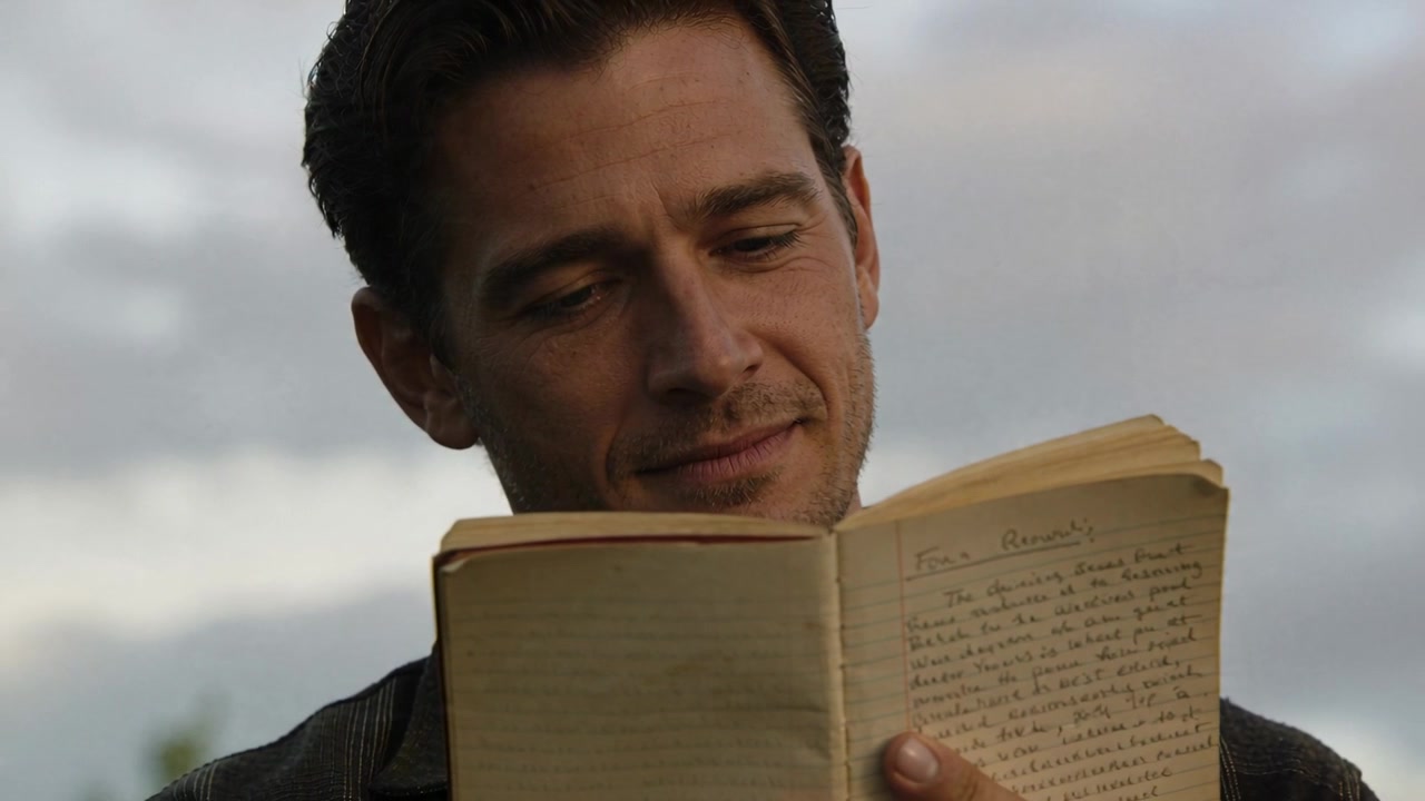 Um homem vestindo camisa de gola escura abre um caderno gasto, lendo anotações ao ar livre, relembrando memórias.