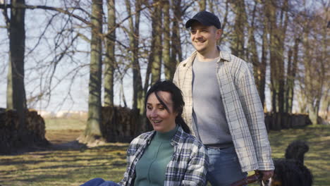 caucasian man carrying his partner who has the arms opened on a wheelbarrow in the countryside. their dog walks around them