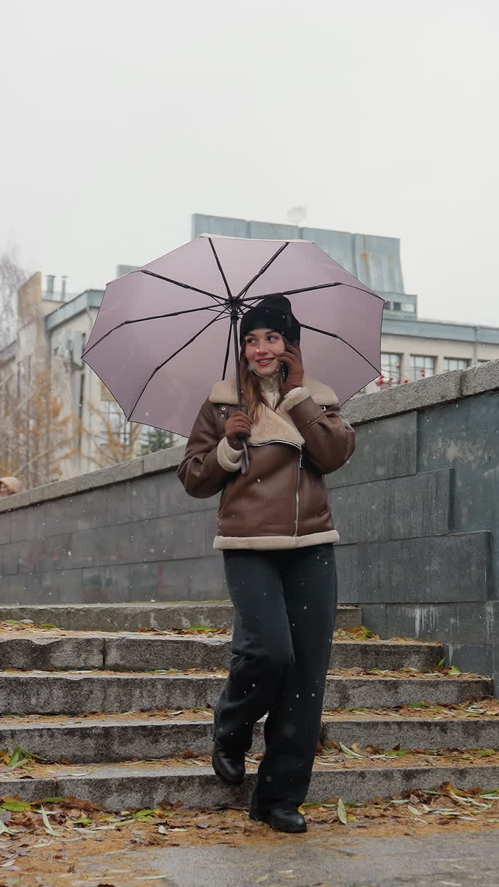 Donna sorridente al telefono che tiene un ombrello, indossa un berretto di lana, una giacca shearling marrone e pantaloni neri, mentre scende dei gradini di pietra durante una leggera nevicata con foglie autunnali colorate