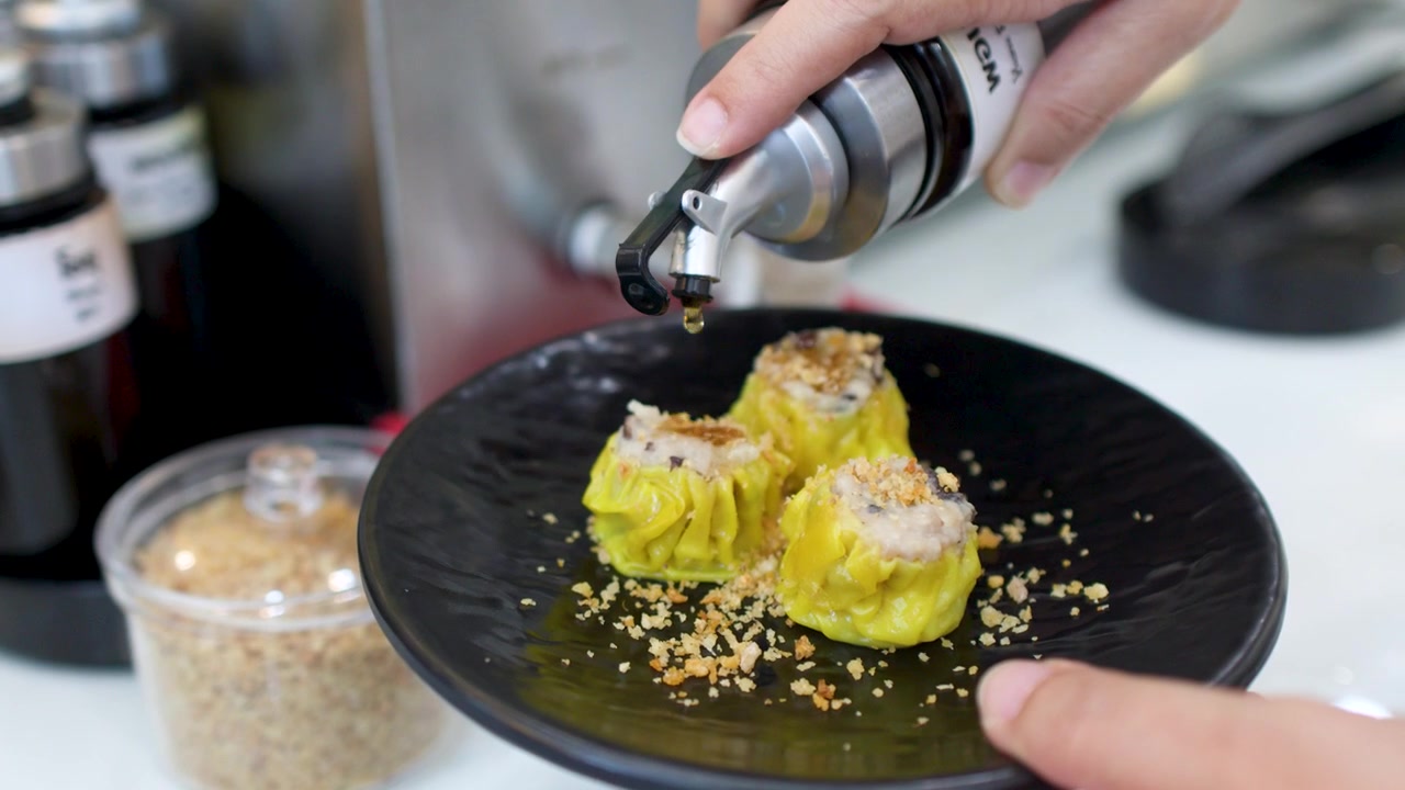Nærbillede af hænder, der hælder mørk sauce over tre gule dim sum-dumplings i Bangkok.
