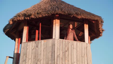 young caucasian woman standing in wooden thatched roof lookout tower takes selfie on cloudless sunny day, handheld pan portrait
