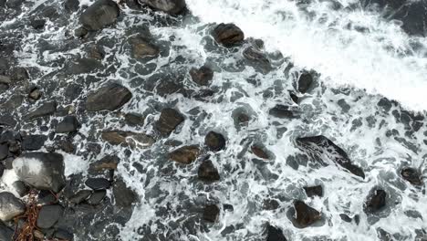 ocean waves splashing up on rocky icelandic beach - aerial starting from birdseye view, slowly tilting up to reveal full north atlantic ocean