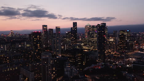 Ascending-footage-of-skyscrapers-in-Canary-Wharf-business-centre-at-dusk.-Tall-illuminated-buildings-against-pink-twilight-sky.-London,-UK