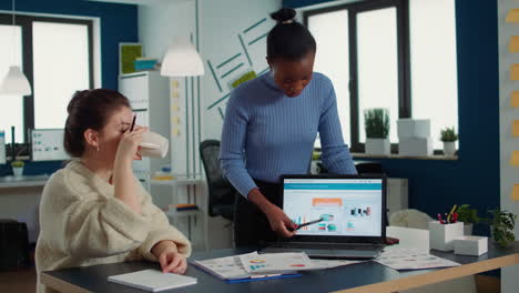 african american woman presenting sales charts using laptop to coworker drinking coffee or tea