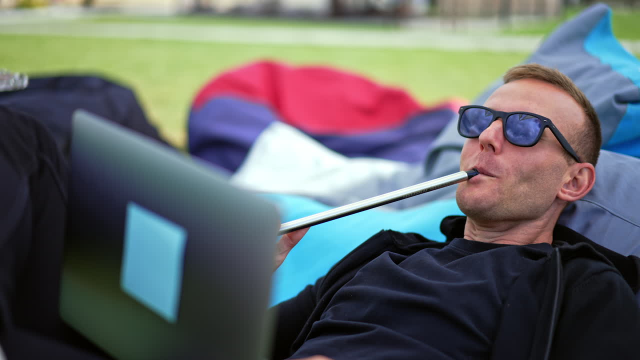 Smiling male freelancer working on computer combining it with rest outdoors. Man smoking hookah producing thick smoke.
